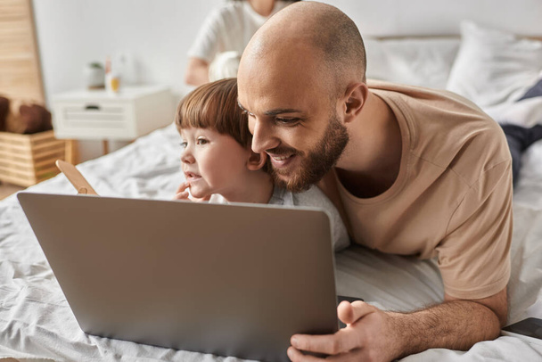feliz padre barbudo abrazando a su pequeño hijo y mirando alegremente a la computadora portátil, concepto de familia - Foto, Imagen