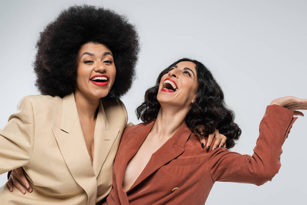 aufgeregte multirassische Frau lacht neben afrikanisch-amerikanischer Freundin über graue, bunte Anzüge - Foto, Bild