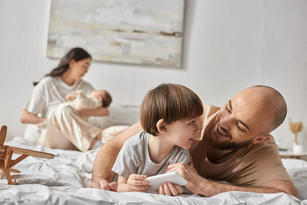 freudig liebender Vater, der seinen kleinen Sohn anlächelt, während er neben seiner verschwommenen Frau und seinem Baby im Bett liegt - Foto, Bild