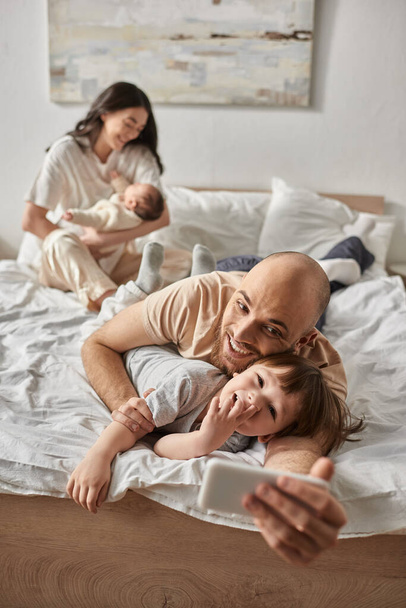 Vertikale Aufnahme eines liebevollen Vaters, der Selfies mit seinem kleinen Sohn neben seiner verschwommenen Frau und seinem Baby macht - Foto, Bild