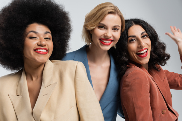 vidám multiracionális nő integet kezét közelében mosolygós női barátok színes öltönyben szürke - Fotó, kép