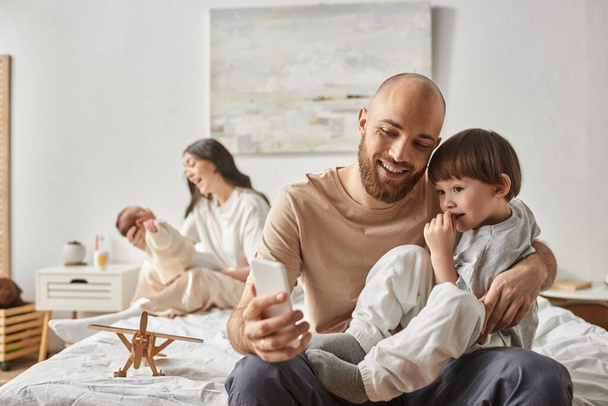幸せな父親と彼の息子に焦点を当てる 彼のぼやけた妻と赤ちゃんと一緒にセルフィーを取る 背景,家族 - 写真・画像