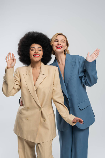 glimlachende multi-etnische vrouwelijke vrienden in veelkleurige pakken zwaaien handen op grijs, fashion shoot - Foto, afbeelding