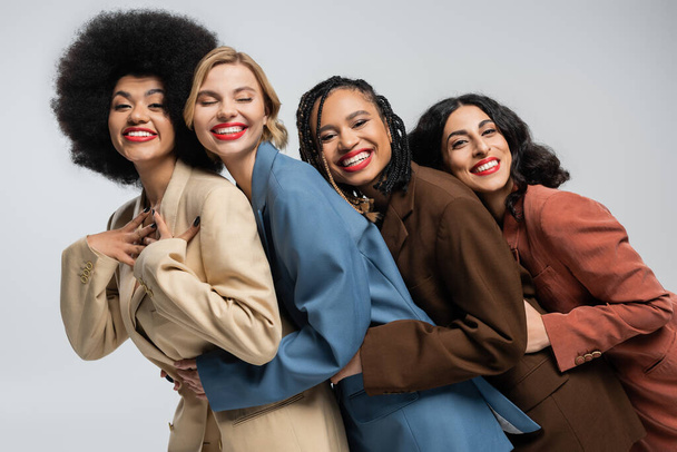 Gruppe trendiger multiethnischer Freundinnen in bunten Anzügen, die sich in grau umarmen und in die Kamera lächeln - Foto, Bild