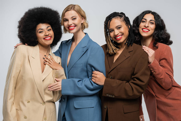 vier vrolijke multiraciale vriendinnen in trendy kleurrijke pakken die glimlachen op de camera op grijs, diversiteit - Foto, afbeelding