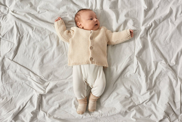 vista superior del adorable bebé recién nacido niño en cárdigan beige cálido acostado sobre una manta blanca con los brazos levantados - Foto, imagen
