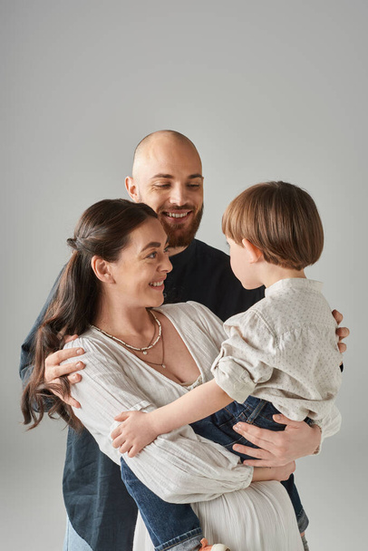 joyous attractive parents looking lovingly at their preschool son on gray backdrop, family - Foto, Imagen