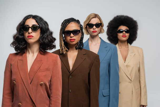 groupe diversifié de copines multiraciales à la mode dans des lunettes de soleil sombres et des costumes colorés sur gris - Photo, image