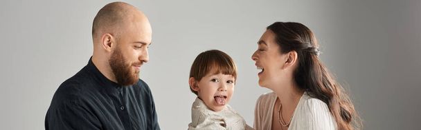 игривый маленький мальчик высовывает язык перед камерой, в то время как в руках его счастливых родителей, баннер - Фото, изображение