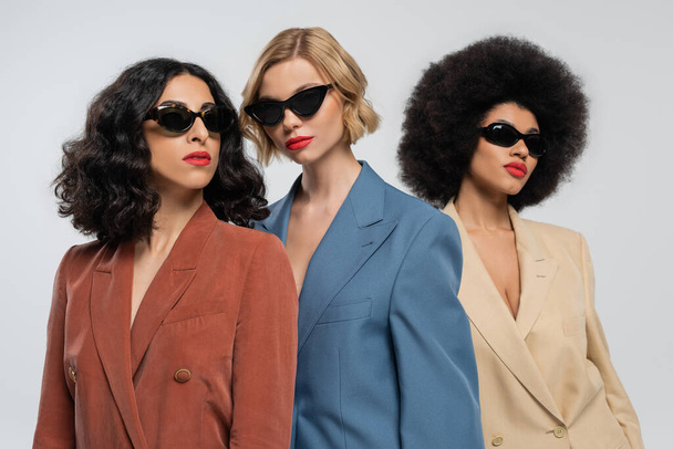 tre ragazze multirazziali alla moda in abiti multicolori e occhiali da sole scuri su grigio - Foto, immagini