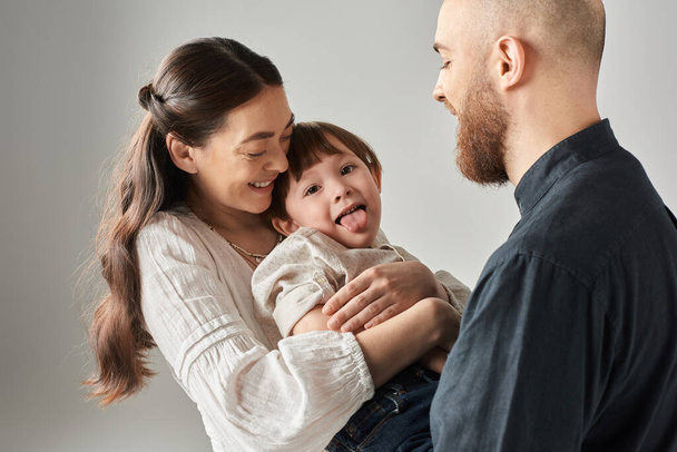 šťastní rodiče ve stylovém oblečení drží svého hravého syna, který vystrkuje jazyk před kamerou - Fotografie, Obrázek