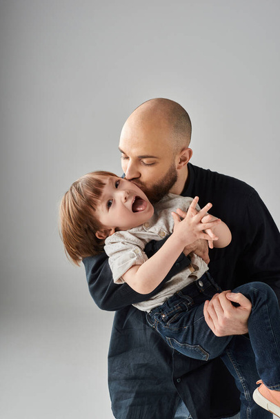vertical shot of bearded father kissing his joyful happy little son in cheek, modern parenting - Photo, Image