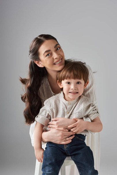 colpo verticale di madre gioiosa che abbraccia il suo piccolo figlio sorridente su sfondo grigio, genitorialità moderna - Foto, immagini