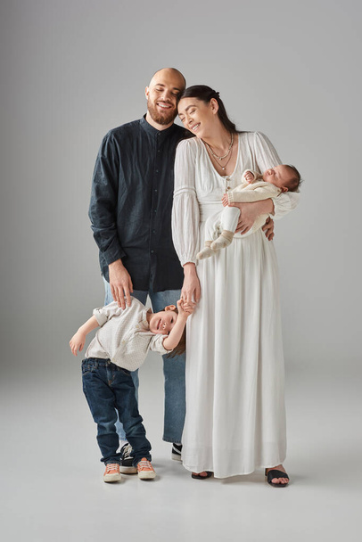 šťastný moderní rodiče v nóbl oblečení s úsměvem a pózování se svými dětmi na šedém pozadí - Fotografie, Obrázek
