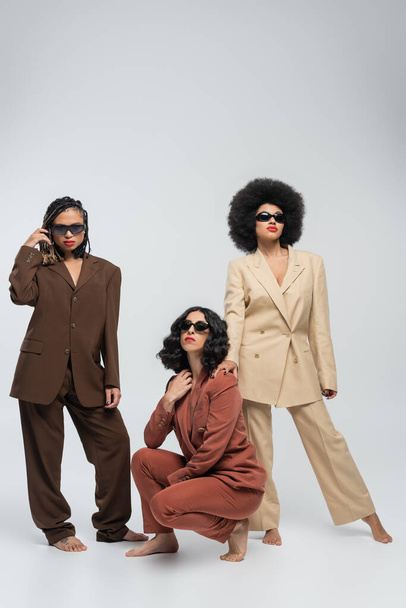 barefoot multiracial female model in trendy colorful suits and dark sunglasses on grey, full length - Foto, Imagem