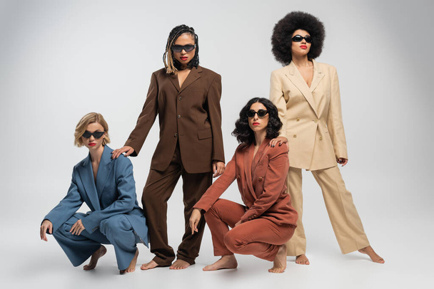 різноманітна група босоніж і стильні багатоетнічні подружки в костюмах і темних сонцезахисних окулярах на сірому - Фото, зображення
