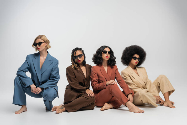 diverse fashion, barefoot multiethnic girlfriends in sunglasses and colorful suits sitting on grey - Photo, Image