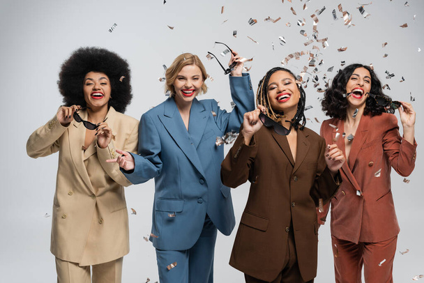 trendy multiracial girlfriends in colorful suits rejoicing under falling confetti, party time - Photo, Image