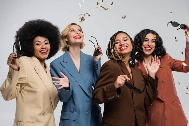 trendige multikulturelle Freundinnen in bunten Anzügen lächelnd unter fallendem Konfetti, Party Time - Foto, Bild
