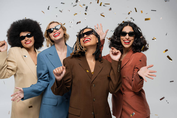 dolblij multiraciale vriendinnen in zonnebrillen en trendy pakken onder kleurrijke confetti op grijs - Foto, afbeelding