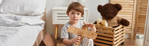 Sevimli, küçük bir çocuk ev elbisesi içinde tahta uçak oyuncağını tutuyor ve kameraya bakıyor, afiş yapıyor. - Fotoğraf, Görsel