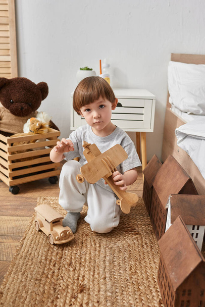 vertical shot of adorable little boy playing actively with wooden plane and looking at camera - Foto, imagen