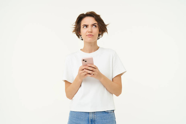 Portret myślącej kobiety ze smartfonem, wyglądającej na zmieszaną, zastanawiającej się, podejmującej decyzję, stojącej na białym tle studia. Przestrzeń kopiowania - Zdjęcie, obraz