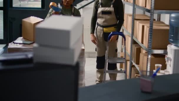 Manager wearing protective equipment in warehouse supervising african american trainee sealing cardboard box packages. Storage area personnel properly securing goods to avoid problems during shipping - Footage, Video