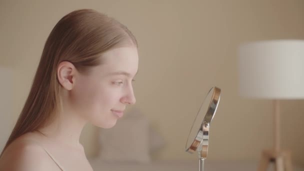 Ικανοποιημένο ελκυστική νεαρή κοπέλα κοιτάζει στον καθρέφτη αντανάκλασης, απολαμβάνοντας αποτέλεσμα μετά την εφαρμογή του καλλυντικού προϊόντος. Λευκή νεαρή γυναίκα με τέλειο δέρμα. Διαφήμιση φροντίδας δέρματος. Αργή κίνηση - Πλάνα, βίντεο