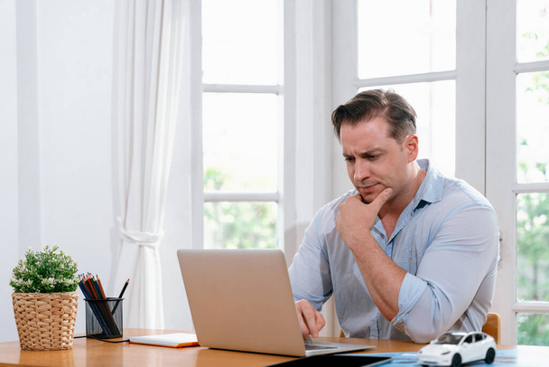 Geschäftsmann müht sich, das Arbeitsproblem von zu Hause aus mit dem Laptop zu lösen, sitzt zu Hause mit gestresstem und frustriertem Gesichtsausdruck auf seinem Schreibtisch und versucht, eine Lösung für das Problem zu finden. Synchronos - Foto, Bild