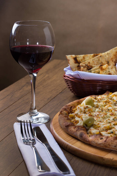 カタピルチキン風味のピザは木製のボードで提供されました. モッツァレラ,チキン,クリームチーズ,グリーンオリーブ,トウモロコシ,赤トマトソースで作られています.赤ワインとコロッティーニのガラス. 垂直写真. - 写真・画像