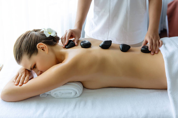 Hot stone massage in spa salon in luxe resort met daglicht sereniteit ambient, gelukzalige vrouw klant genieten spa basalt steen massage glijden over het lichaam met rustgevende warmte. Stil maar. - Foto, afbeelding