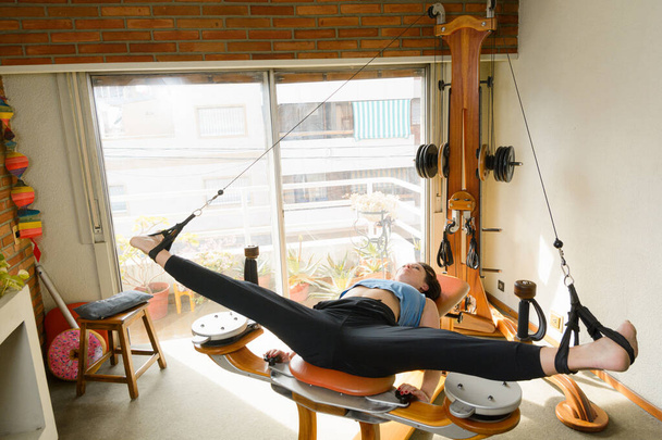 Caucasian woman at home in blue sportswear, lying on her back on exercise machine with pulleys and weight, with legs open training hip joint, next to window in morning before breakfast - Photo, Image