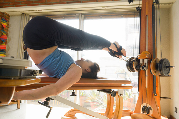 Caucasian woman with blue sports clothing and black hair, at home lying on exercise machine with pulleys and weights, with her curved legs working and elongating joints of vertebrae - Photo, Image
