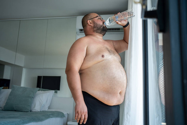 Fat man suffers dying from heat. Bearded obese guy drinks lot water standing at open window next to air conditioner trying to cope with abnormal summer warmth sweats heavily replenishes water balance - Photo, Image