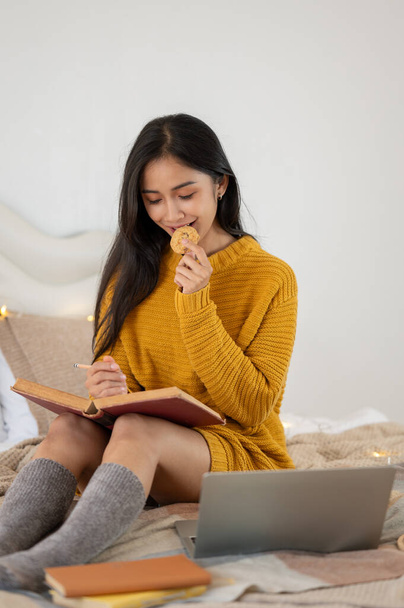 Un ritratto di una donna asiatica felice e bella in un accogliente maglione a maglia che mangia un biscotto mentre legge un libro o tiene il suo diario sul suo letto nella sua camera da letto. - Foto, immagini