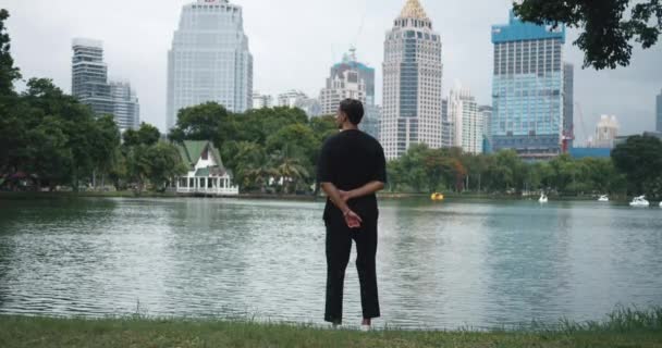 Widok z tyłu 30s mężczyzna stoi ciesząc się patrząc na jezioro w parku miejskim z nowoczesnymi drapaczami chmur budynków w mieście na tle. Widok z tyłu Mężczyzna turysta w pięknym parku w centrum nowoczesnej metropolii - Materiał filmowy, wideo