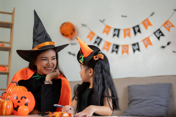 Una mamá asiática alegre y una pequeña hija linda están haciendo decoraciones de artesanía de Halloween y divirtiéndose mientras se preparan para la temporada de Halloween en casa juntos. - Foto, imagen