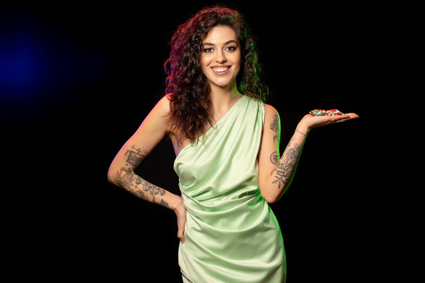 Gambling happy excited young brunette with tattoos on arms standing against black background holding pile of betting chips on palm, cropped shot - Photo, Image