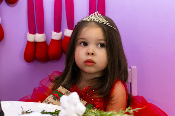 赤い服を着た少女が贈り物を抱いている. 小さな女の子が椅子にプレゼントを持っています. 赤いドレスの子供がギフトボックスを持っています. 幸せな新を祝う多くのギフトボックスを持っているカメラを見ている少女 - 写真・画像