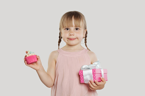 Cute cheerful child girl holding cupcake and pink gift box with white silky ribbon on white background. Happy birthday party concept - Photo, Image