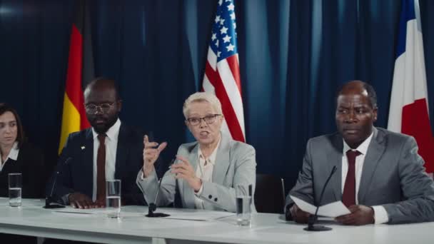 Uluslararası konferansta konuşan ve doğru kelimeleri bulmak için tökezleyen beyaz kadın politikacı, dinleyen çeşitli meslektaşlar ve arka planda ABD, Alman ve Fransız bayrakları - Video, Çekim