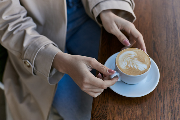cropped view of young woman in trench coat holding cup with cappuccino in outdoor cafe, latte art - Photo, Image