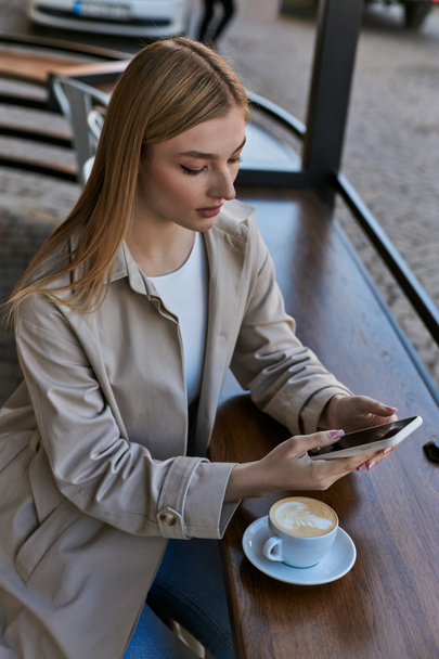blonde young woman in trench coat using smartphone near cup with cappuccino in outdoor cafe - Photo, Image