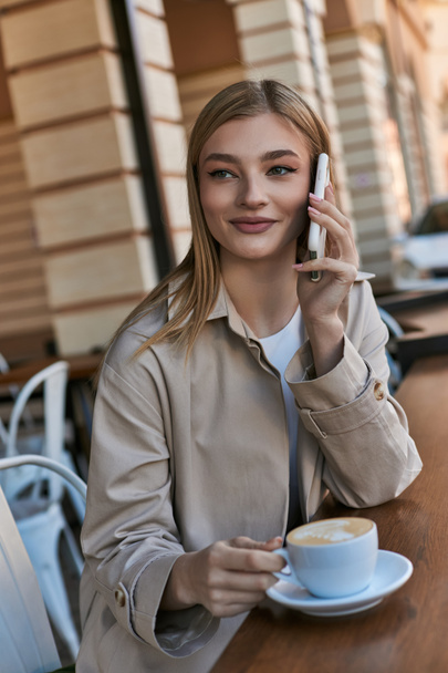 mujer joven feliz en gabardina tener llamada en el teléfono inteligente cerca de la taza con capuchino en la cafetería al aire libre - Foto, imagen