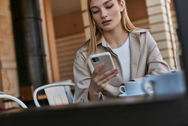 blondýny mladá žena v trench kabát pomocí smartphone v blízkosti šálek s kávou v venkovní terase v kavárně - Fotografie, Obrázek