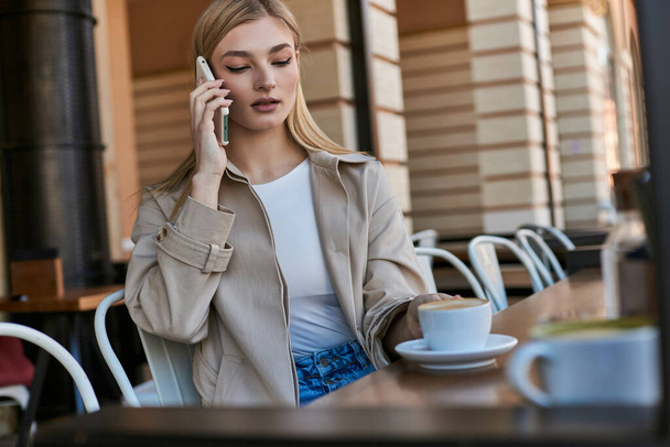 mooie jonge vrouw in trench jas met oproep op smartphone in de buurt van kopje met cappuccino in outdoor cafe - Foto, afbeelding
