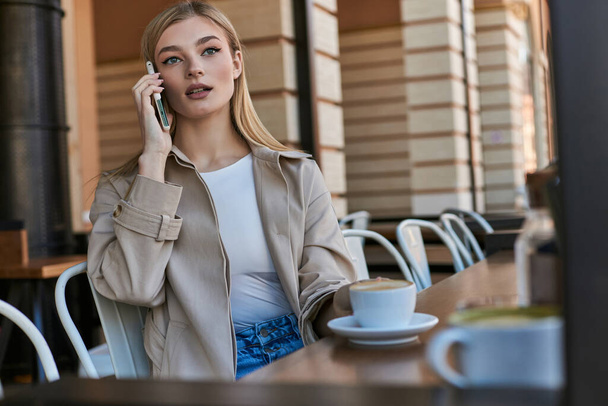 jolie femme en trench coat ayant appel sur smartphone près de tasse avec cappuccino dans un café en plein air - Photo, image