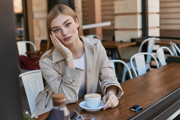 attractive young woman in trench coat enjoying her cup of cappuccino outdoors in cafe, weekend - Photo, Image