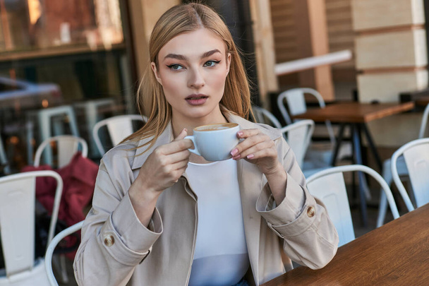 dreamy blonde woman in trench coat enjoying her cup of cappuccino while sitting outdoors in cafe - Photo, Image